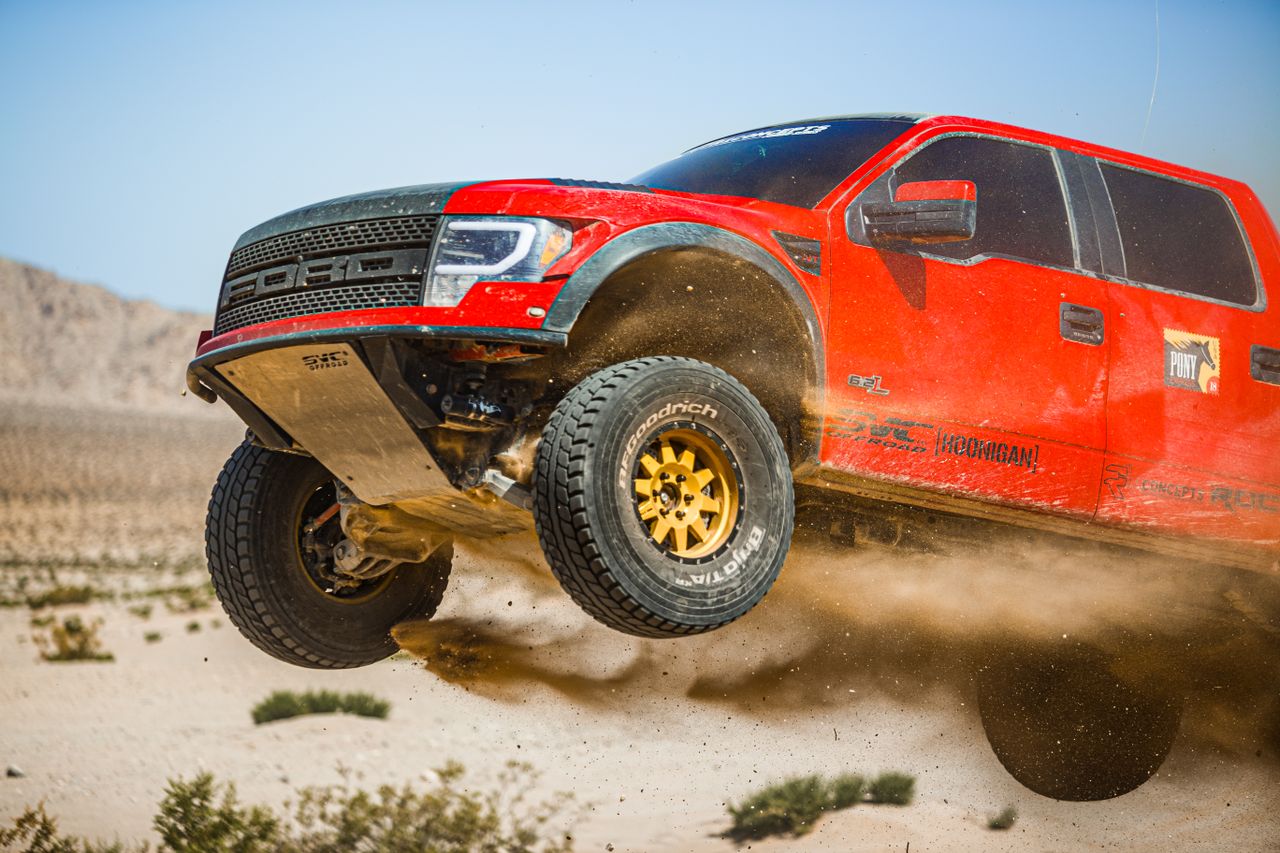 A dynamic shot of a red truck off-roading in the desert, captured mid-air on a sunny day.